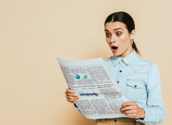 shocked brunette woman in denim shirt reading business newspaper isolated on beige