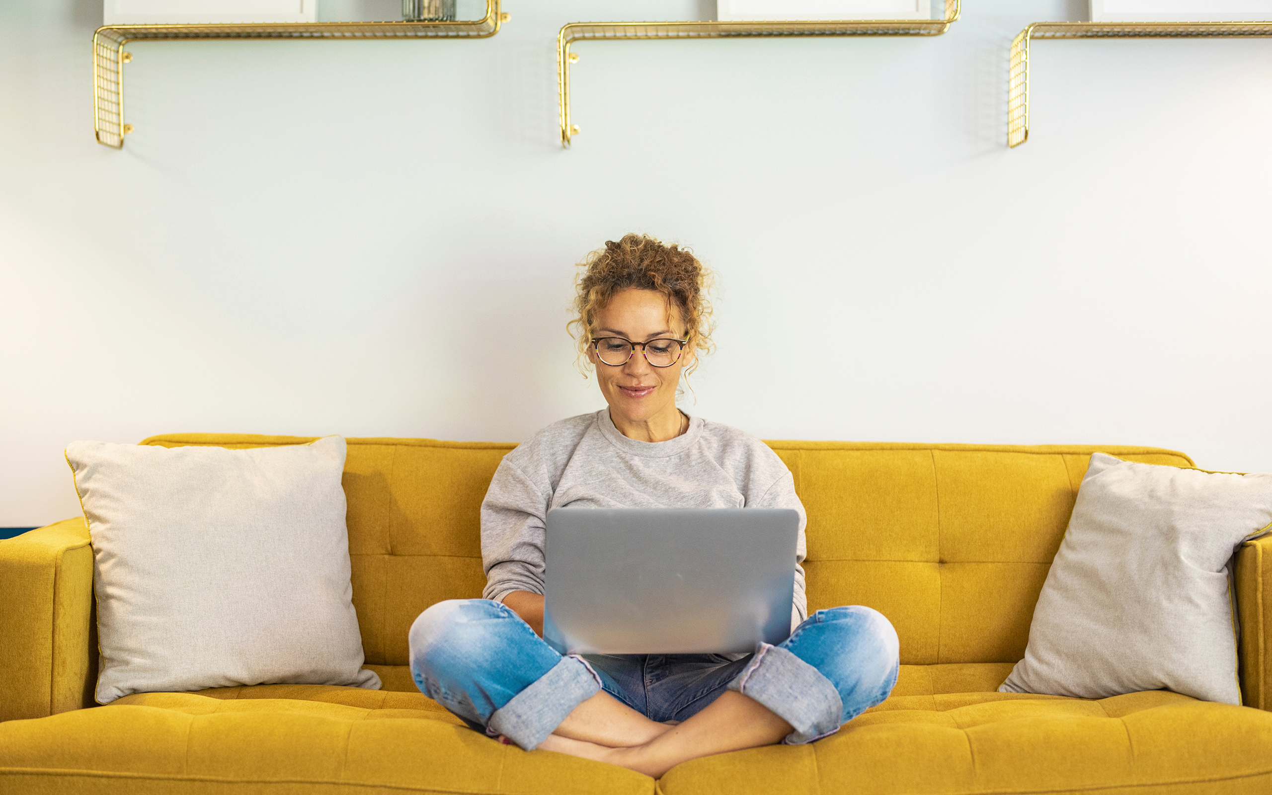 Women sitting cross legged on a mustard colored sofa with a laptop on her lap.
